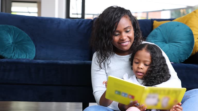 Happy African American mother and daughter reading a book at home sitting on the floor while enjoying their time together